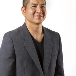 An image of Eric Tang. He is visible from the waist up, and wears a gray blazer over a black shirt. His hair is cut short. He smiles at the camera.