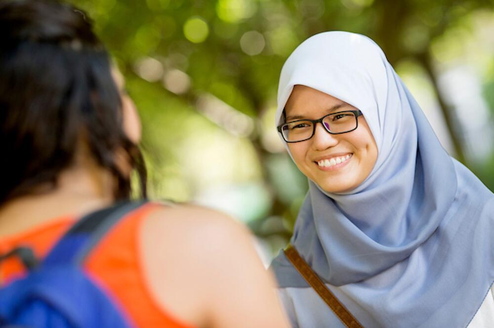 A smiling girl in a light blue hijab greets her friend.